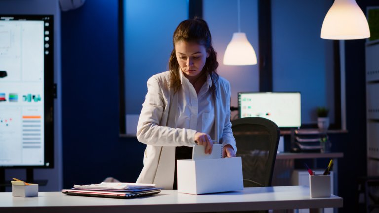 A woman in a white lab coat is packing items into a white box on a desk, as part of her resignation process. The office setting features computer monitors, shelves, and lamps, creating a professional workspace illuminated with blue and white lights.