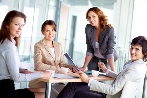 Four women in business attire are gathered around a table in a modern office setting, smiling and engaging in discussion. They have documents and coffee cups on the table, suggesting a professional meeting or brainstorming session that is central to their careers. Light streams in from large windows.
