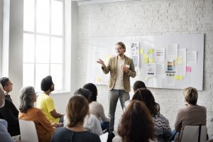 A person gives a training presentation to a seated audience in a bright room. They stand in front of a whiteboard covered with various charts, notes, and papers. The audience, diverse in age and appearance, listens attentively as light pours in through a large window.