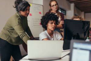 A group of four women developers collaborate in an office setting, with two looking at a desktop computer and another focused on a laptop. They appear engaged and are likely discussing a project. A whiteboard with notes is visible in the background.