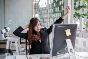 A woman in a black blazer stretches her arms to combat dead butt while seated at her office desk in front of a computer monitor. The workspace features large windows with a geometric pattern and empty chairs in the background. The desk has sticky notes on the monitor and papers scattered around.