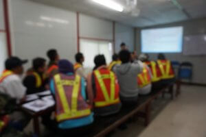 A group of people wearing safety vests sit on benches in a classroom setting, focused on a training session led by a presenter at the front of the room. Information is displayed on a screen, and the room features a whiteboard and fluorescent lighting.