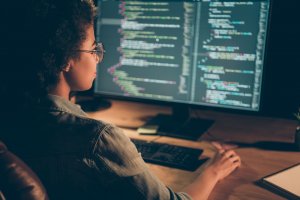 A developer wearing glasses and a denim jacket sits at a desk, working on a computer with large monitors displaying colorful lines of code. The dimly lit setting suggests an intense focus on programming or software development.