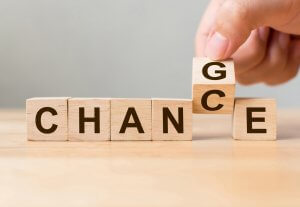 A hand is exhibiting leadership by rearranging wooden blocks that spell "CHANGE" to instead spell "CHANCE" by replacing the "G" block with a "C" block. The blocks rest on a wooden surface against a pale background.