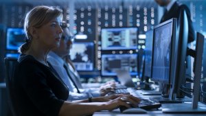 A woman working at a computer with multiple screens in a dimly lit office. Two colleagues are sitting beside her, also focused on their monitors. The background shows a tech-heavy environment with several monitors displaying data and network visuals, reminiscent of control rooms used by governments.