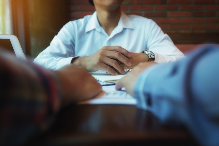 Three people at a meeting table, with one person in the center facing forward and the other two barely in the frame. The central person, likely leading the negotiation, is wearing a white shirt and has hands clasped on the table. Blurred papers and a laptop are visible in the foreground.