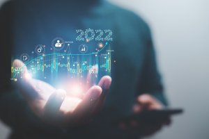 A person holds a holographic projection featuring graphs, charts, and various icons related to data and technology. The number "2022" is prominently displayed above the data-driven projection, signifying a focus on trends or developments in 2022.