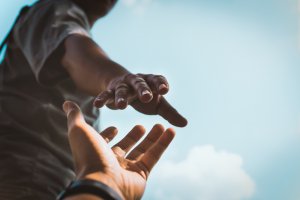 A person is reaching down with their hand, and another hand from below is reaching up towards them. The background is a clear blue sky with a few clouds, creating a hopeful and humanitarian atmosphere.
