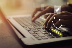 Hands typing on a laptop keyboard with superimposed digital icons, including a check mark, a smiley face, and a row of five stars, indicating positive feedback or a rating system. Light filters softly through the background.