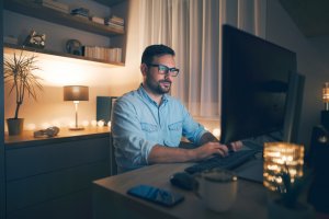 A man is sitting at a desk in a dimly lit room, remote working on his computer. He is wearing glasses and a blue shirt. A smartphone, coffee mug, and candles are on the desk. Shelves with books and decorative items are in the background, and a window is partly covered by a curtain.