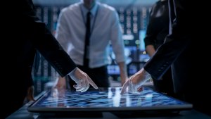 A group of people in business attire are gathered around a large interactive touchscreen table displaying complex data. Two individuals are pointing at the screen, which showcases intricate graphics. The setting appears to be a modern conference room or control center.