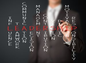 A person in a suit is writing words related to leadership on a transparent board. The central word "LEADERSHIP" is highlighted in red, surrounded by terms like "communication," "teamwork," "influence," and more.