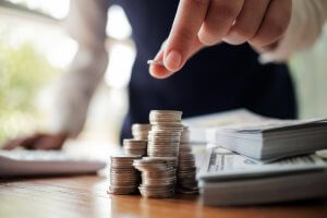 A person is stacking coins on a wooden table with one hand, while the other hand reaches for a coin to pay. A calculator and stacks of paper money are in the background, suggesting financial planning or budgeting.