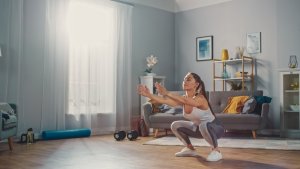 A woman in athletic wear performs a squat exercise in her bright, modern living room, which doubles as a home gym with a sofa, bookshelf, and fitness equipment like a yoga mat and dumbbells. Sunlight streams through the window, creating a warm and energized atmosphere.