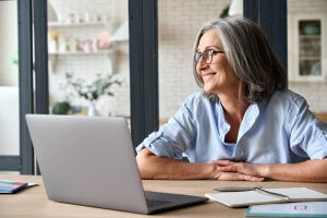 A smiling elderly woman with gray hair and glasses sits at a desk in front of a laptop, perhaps navigating new career opportunities. She wears a light blue shirt and looks to the side. There are papers, a pen, and a phone on the desk, with shelves and a brick wall in the background.