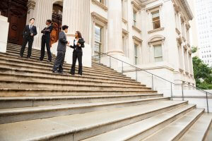 Four business professionals, three men and one woman, stand and converse on the steps of a grand, neoclassical government building. They are dressed in formal business attire and hold documents in their hands. The building features tall columns and a large doorway.