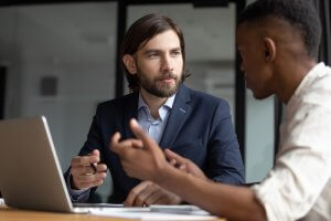 A bearded man in a suit sits at a desk with an open laptop, engaged in a discussion about pay with another person gesturing with their hands. Both are seated indoors, possibly in an office environment. The scene appears to depict a professional conversation or meeting.