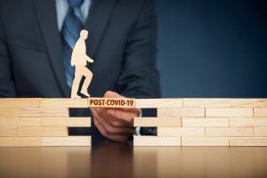 A person in a suit holds a wooden plank labeled "POST-COVID-19" that bridges a gap between two stacks of wooden blocks. A wooden cutout of a walking figure is placed on the plank, symbolizing progression beyond the COVID-19 pandemic and the return to work.