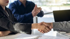 Two people in business attire shaking hands across a table, signifying an agreement or deal. Another person in the background is clapping, and documents related to a government job are visible on the table. A laptop is also partially visible.