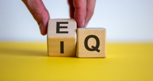 A hand is holding two wooden blocks, one with the letter "E" and the other with "Q," representing EQ, placed beside each other on a yellow surface. The background is white.