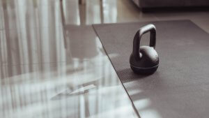 A black kettlebell sits on a gray exercise mat in a home gym with a shiny floor. Sunlight streams through vertical blinds, casting shadows on the floor and the kettlebell.