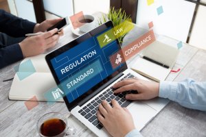 A person works on a laptop displaying words: Regulation, Standard, Law, Compliance. Another person is using a smartphone. A coffee cup, notepad, and pen are on the desk, underscoring the professional office setting focused on compliance.
