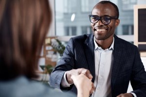 A man wearing glasses and a formal suit smiles as he shakes hands with another person in an office setting. The background shows office furniture and a large window, suggesting a professional environment. The man's expression reflects positivity and friendliness, possibly celebrating his new government job.