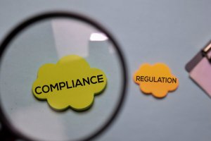 A magnifying glass focuses on a green cloud-shaped paper with the word "COMPLIANCE" prominently displayed, while a yellow cloud-shaped paper labeled "REGULATION" lies nearby on a light blue background.