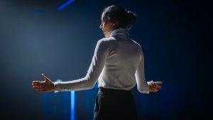 A woman in a white turtleneck and black pants stands on a dimly lit stage, speaking passionately with her hands extended. Her hair is tied back, and she is wearing a headset microphone. Blue lights illuminate the background, creating a dramatic atmosphere that highlights the power of women.