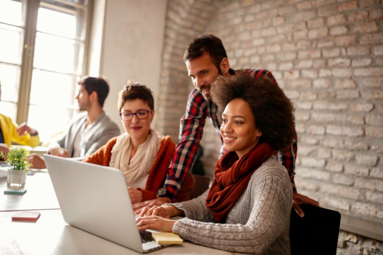 A diverse group of four adults collaborating in a bright office with brick walls exemplifies a positive work culture. Two women and a man are gathered around a laptop, smiling and engaged, while another person in the background speaks with a colleague. A small plant is on the table.