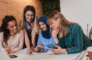Four women are gathered around a table in the workplace, smiling and collaborating on a project. They are engaged and pointing at documents spread out on the table. One woman is holding a phone, another is writing, and the atmosphere appears friendly and productive.
