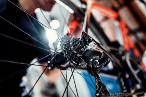 Close-up view of a bicycle's rear gear system and chain. A person is adjusting or inspecting the bike, likely working towards their cycling goals. The image focuses on the intricate details of the gears, chain, and derailleur with the bicycle wheel spokes visible in the foreground.