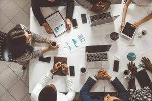 A diverse group of people sit around a table engaged in a meeting. Laptops, tablets, smartphones, coffee mugs, papers with graphs, and notebooks are spread out on the table. Their hands are either resting on the table, holding coffee, or using electronic devices to enhance communication.