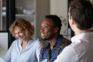 Three people are engaged in a conversation or a meeting. One person, wearing a plaid shirt, exudes leadership while speaking, and the others, dressed casually, are smiling and attentive. The background shows a modern office setting with shelves and blurred items.