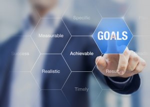 A person in a suit points to a hexagon with the word "GOALS" on a transparent virtual interface. Surrounding hexagons contain words like "Specific," "Measurable," "Achievable," "Realistic," and "Timely," underscoring essential goal-setting concepts aimed at success.