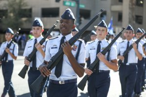 A group of young uniformed ROTC cadets march in formation during a parade. They are dressed in blue trousers and white shirts with ties, carrying rifles on their shoulders. The background includes buildings and a green street sign.