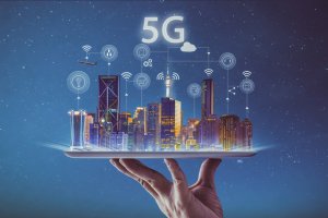 An illuminated cityscape with various technological icons representing connectivity is shown floating above an outstretched hand. The background features a starry night sky with the term "5G" prominently displayed above the city, emphasizing its role in transforming urban connectivity.