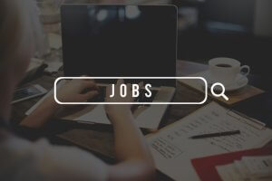A person types on a laptop at a desk with a coffee cup, papers, and a pen nearby. The word "JOBS" is prominently displayed in a large search bar overlay, emphasizing an online job search for government positions. The background is blurred, focusing on the search prompt.