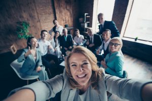 A woman in the foreground takes a group selfie with ten smiling colleagues in a well-lit workplace. They are all giving a thumbs-up or smiling at the camera, suggesting a positive and cheerful atmosphere. The room has wooden walls and large windows.