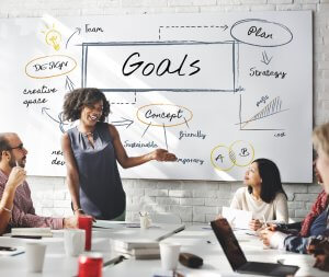 A group of people in a meeting room discussing goals. A woman is standing and presenting in front of a whiteboard with words like "design," "strategy," "concept," and "plan" written on it. Two people are seated at a table with laptops and documents, focusing intently on achieving their goals.