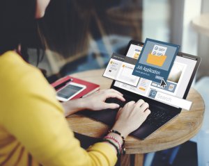 A person wearing a yellow shirt is typing on a laptop at a round wooden table. The screen displays a "Job Application" interface with a search bar, possibly for government job listings. To the left of the laptop, a closed red notebook and a pen are placed on the table.