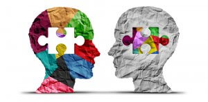 Two crumpled paper silhouettes of human heads face each other. The left head is colorful and has a white puzzle piece cut out, while the right head is grayscale with a multicolored puzzle piece inserted in the cutout, symbolizing connection and understanding across different cultures.