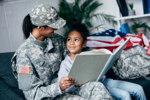 A military personnel in uniform, possibly from ROTC, sits beside a young child on a couch, holding a large book. The child looks up at the military personnel with a smile. An American flag and military gear are visible in the background.