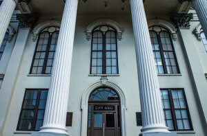 A front view of a historic government building with tall white columns and arched windows. The central door has a sign above it reading "CITY HALL." The architecture features decorative molding and classic elements.