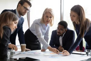 A group of five diverse business professionals is gathered around a table in the bustling workplace, intently discussing and pointing at documents and sticky notes. The environment appears collaborative, with a focus on teamwork and planning.