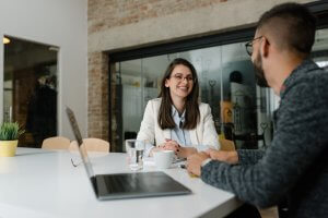 A woman and a man are sitting at a white table in an office, engaged in a conversation about career advice. The woman is smiling while wearing a white blazer and glasses. The man has a beard and is wearing a gray sweater. A laptop, coffee cups, and a plant are on the table.