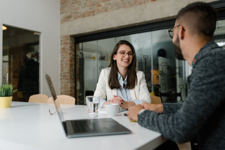 A woman and a man are sitting at a white table in an office, engaged in a conversation about career advice. The woman is smiling while wearing a white blazer and glasses. The man has a beard and is wearing a gray sweater. A laptop, coffee cups, and a plant are on the table.