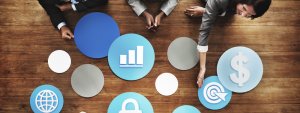 Three people are arranging round icons on a wooden table, symbolizing concepts like a globe, bar chart, lock, target with an arrow, and a dollar sign. The individuals appear focused on organizing these symbols, suggesting a collaborative planning effort crucial for career growth.