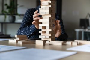 A person wearing a dark blue polka-dotted shirt is carefully removing a block from a tall Jenga tower on a wooden table in the workplace. There are several blocks scattered around the base of the tower, with indoor plants and blurred office items in the background.