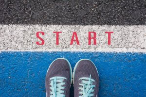 Close-up of a pair of gray athletic shoes with teal accents positioned at a starting line on a blue running track, symbolizing the beginning of your journey. The word "START" is written in red letters on the white section of the track. The top half of the image shows the black asphalt track. Use these steps as career advice to propel yourself forward.